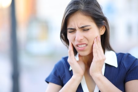 Stressed woman outdoors, appearing in pain and holding her jaw, showing symptoms of TMJ disorder and potentially seeking TMJ BOTOX treatment
