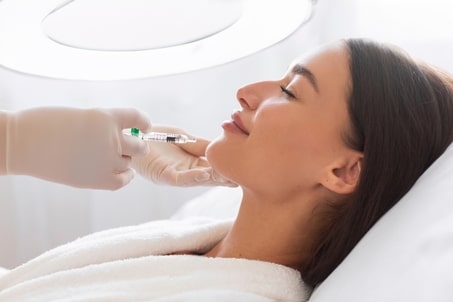 Side view of a cosmetologist administering a Botox injection to a young woman's chin during a skincare session, illustrating the use of dental Botox for facial aesthetics and muscle relaxation