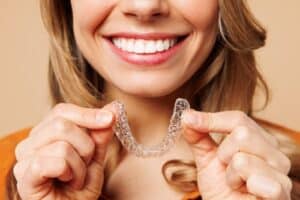 Close-up of a young woman in an orange shirt holding invisible aligners for teeth straightening, showcasing an Invisalign smile and modern lifestyle concept against a light beige background.