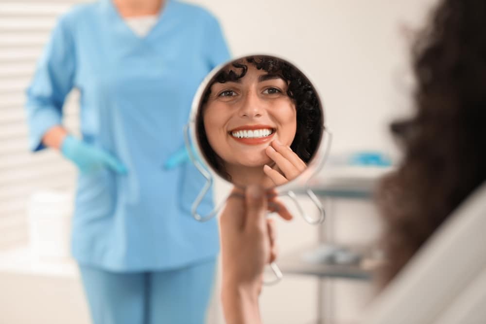 Patient looking in the mirror while a dentist observes in the clinic, showcasing results of dental veneers and crown bridges.