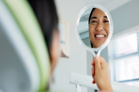 Woman smiling and checking her teeth in a mirror at the dentist, showcasing satisfaction after teeth whitening and routine oral care
