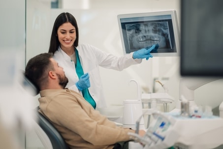 A smiling female dentist explains a full mouth restoration diagnosis by pointing to a dental X-ray on a digital screen in a modern clinic