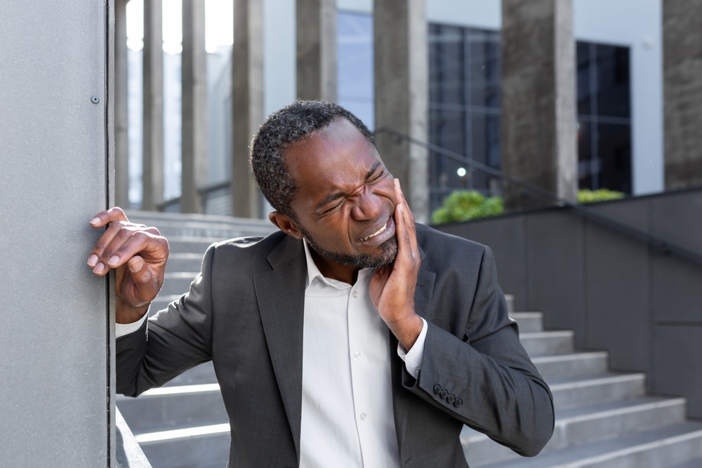 Man in suit standing outside office building holding his cheek in pain, showing toothache and oral discomfort."