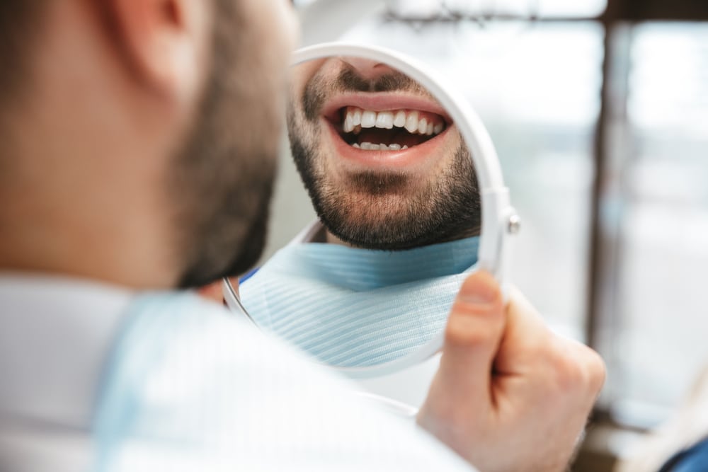 Man smiling while examining his teeth in a mirror after a routine dental check-up and cleaning.