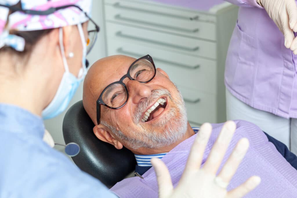 Older man smiling in a dental chair while two dental professionals discuss his care.