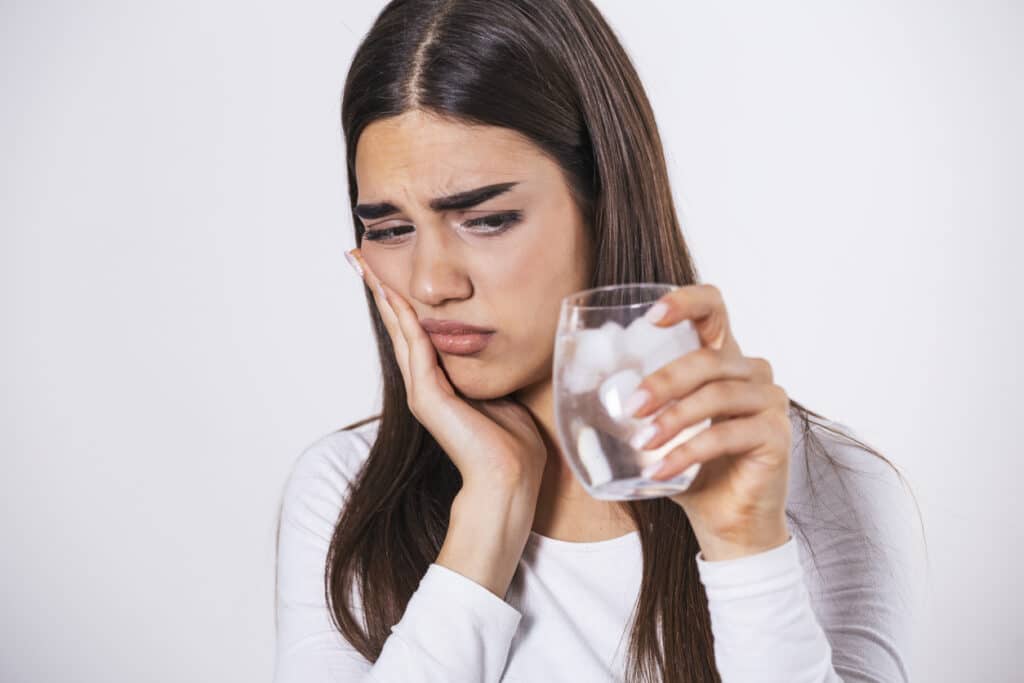 Woman holding a glass of ice water while touching her cheek due to tooth sensitivity.