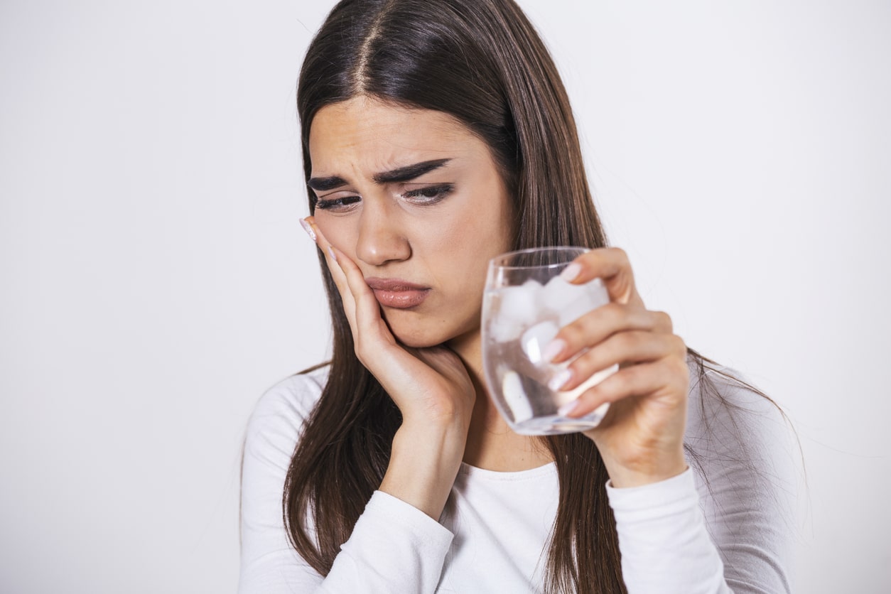 Woman holding a glass of ice water while touching her cheek due to tooth sensitivity.